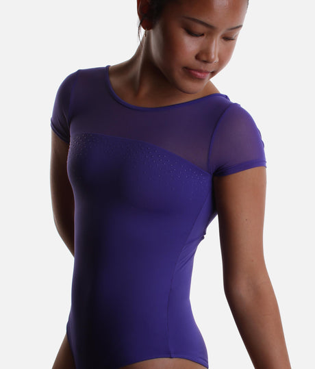 A woman wearing a short-sleeve leotard in a deep purple color, featuring sheer mesh panels across the chest and shoulders, against a plain light gray background.
