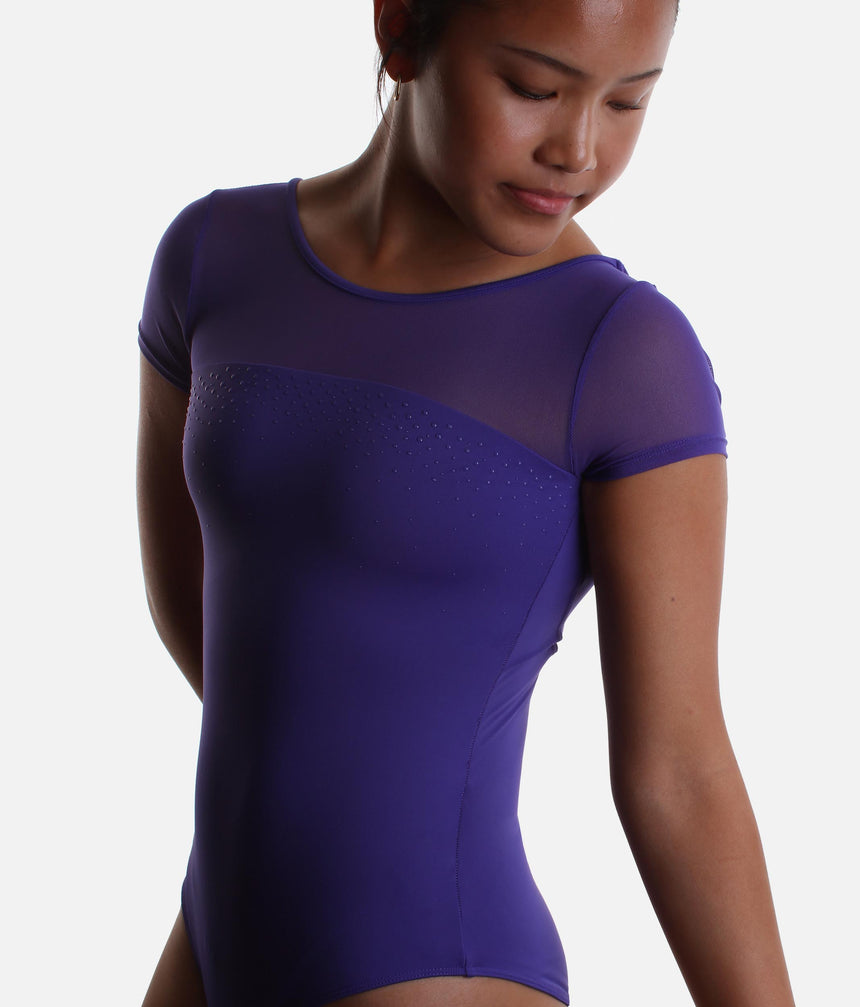 A woman wearing a short-sleeve leotard in a deep purple color, featuring sheer mesh panels across the chest and shoulders, against a plain light gray background.