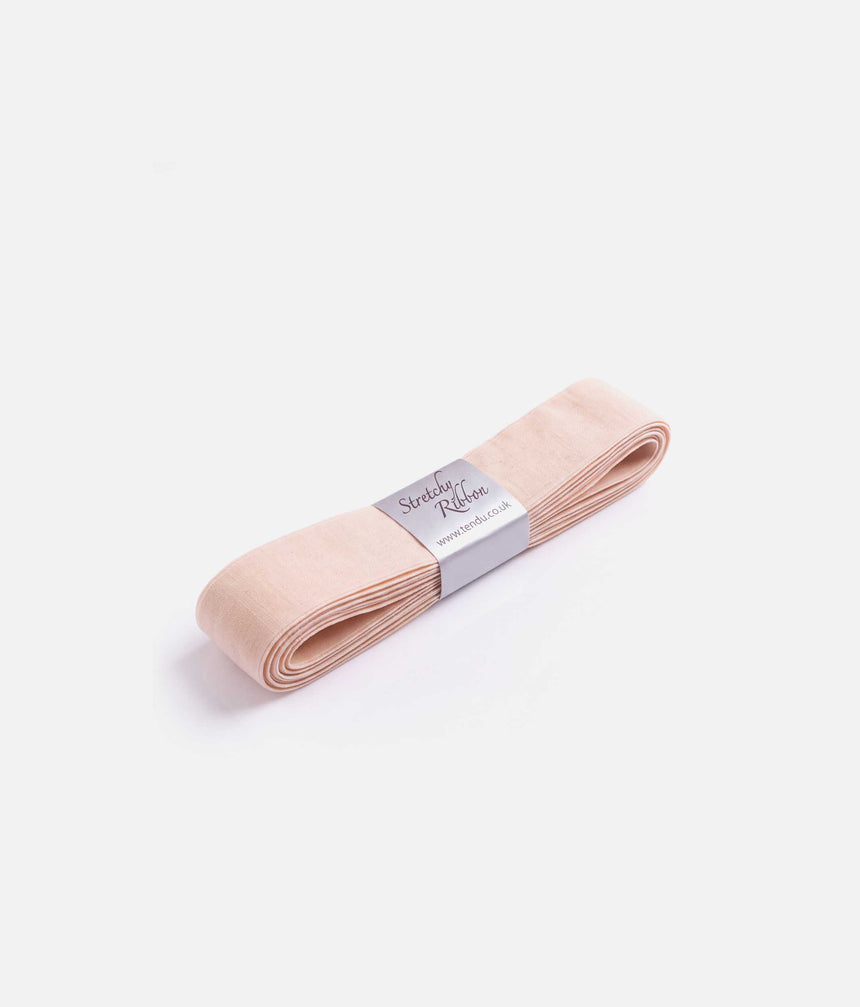 Angled view of folded pale peach-pink Tendu stretchy pointe shoe ribbon with a silver-grey paper label on a white background.